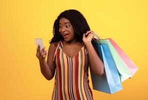 Huge sale in web store. Shocked black lady with shopping bags looking at smartphone, using new mobile app, purchasing things online over orange studio background photo