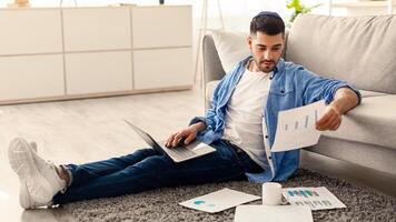 Paperwork Concept. Portrait of young jewish man working at home office, holding and reading paper or financial document, sitting on floor leaning on couch and using laptop computer in living room photo