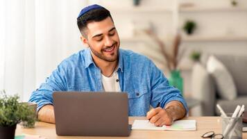 Online Training. Portrait of happy jewish man in kipa working or studying remotely at home office, writing in his notebook with pen, taking notes, using laptop and sitting at table. E-learning photo