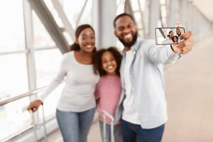 Capturing Moment. Happy loving black family of three taking selfie, posing at airport, hugging and cheerfully smiling at smartphone, selective focus on device gadget screen, blurred background photo