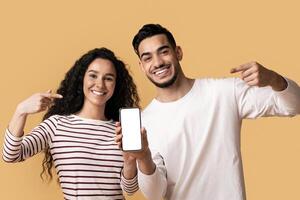 Happy Middle Eastern Couple Showing Blank Smartphone Screen For Mockup, Cheerful Arabic Man And Woman Demonstrating New Mobile App While Standing Together Over Yellow Background, Copy Space photo