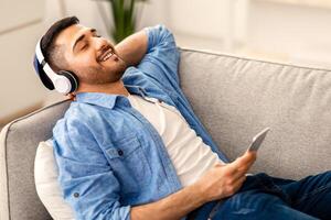 Rest And Relax Concept. Portrait of calm jewish man lying on sofa, listening to music, audio book, podcast, enjoying meditation for sleep and peaceful mind in wireless headphones. Zen-like, banner photo