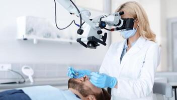 Innovative instruments in modern microdentistry. Female dentist looking at dental microscope at brand new clinic, using innovative tools while making treatment for male patient, side view, copy space photo