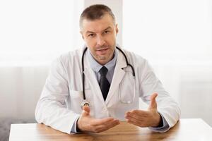 Physician Office Visit Concept. Portrait of male doctor talking to camera, gesturing, consulting patient sitting at desk photo