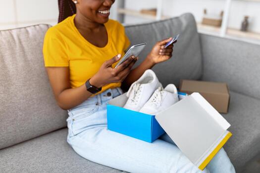 alegre negro mujer desempacar paquete o empaquetar con nuevo Deportes zapatos, participación crédito tarjeta y teléfono inteligente, escritura en línea Tienda comentario, adentro. contento afro dama compras en web, satisfecho con bueno Servicio foto