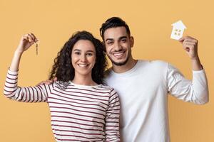 Family Housing. Happy Arab Couple Holding Home Keys And Paper House Icon, Joyful Middle Eastern Spouses Celebrating Moving To Their Own Apartment, Standing Over Yellow Studio Background, Free Space photo