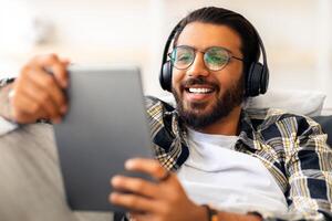 Joyful arab guy watching videos on digital tablet at home, using wireless stereo headset, laying on couch. Happy middle-eastern man relaxing at weekend, watching nice movie online photo