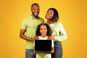 Excited black family of three showing laptop with blank black screen, smiling girl holding gadget with free space for mockup, standing over yellow background and smiling to camera photo