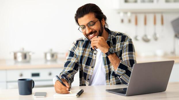 contento Medio este chico independiente contratista sentado en frente de ordenador portátil a cocina, tomando notas, Bebiendo café, mirando para creativo soluciones, indio hombre mirando para trabajo en línea, Copiar espacio foto