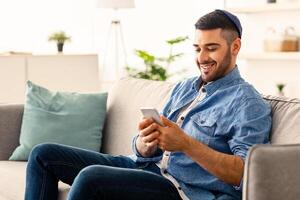 Mobile Communication Concept. Portrait of happy young jewish man using modern smartphone device sitting on comfy sofa at his apartment. Cheerful guy typing sms message, browsing social network photo