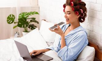Cheerful Lady Using Voice Assistant On Cellphone Working At Laptop Computer Sitting In Bedroom, Curling Hair With Curlers. Distant Work From Home, Freelance, Smart Phone App Concept. photo
