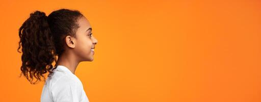 Side view profile portrait of cute African American girl with beaming smile and curly hair in ponytail looking aside at empty free copy space isolated on yellow studio background, banner, panorama photo