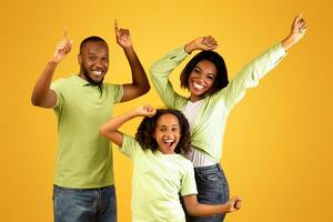 Excited young black family of three dancing and fooling around, enjoying favorite music together on yellow studio background. African american man pointing fingers up at free space photo
