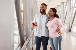 Happy African American Family Of Two People Of Color Waiting For Their Flight In Airport. Smiling Black Couple Hugging Holding Passports With Tickets, Waiting For Departure, Looking Out Of Window photo