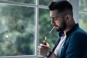 Urban lifestyle, emotional portrait of young handsome man sad and depressed, smoking. Unhappy feeling bittered suffer some problem. Bearded man lights cigarette with lighter, on window background photo