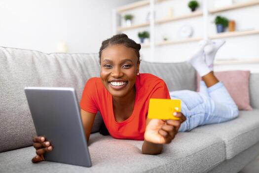 contento africano americano mujer haciendo en línea compra en tableta ordenador personal, utilizando crédito tarjeta, ordenando bienes en web mientras acostado en sofá a hogar. Internet compras, comercio electrónico, remoto bancario concepto foto
