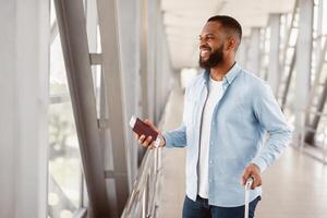 Portrait Of Happy African American Man Waiting For His Flight In Modern Airport. Smiling Black Guy Holding Passport With Ticket, Waiting For Departure, Looking Out Of Window, Free Space photo