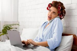 Confused Housewife Talking On Mobile Phone Using Laptop Computer Working Online Sitting In Bed In Modern Bedroom At Home. Mobile Communication, Distance Job And Freelance Work. Side View photo