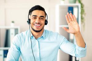 Remote communication concept. Happy Arab guy with headset having call at office, waving at webcam. Head shot of young man using computer for business meeting or web conference photo