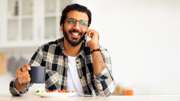 alegre árabe chico teniendo sano almuerzo a hogar, hablando en móvil teléfono y sonriente, de cerca retrato. hermoso indio hombre con jarra de café y plato con ensalada sentado a cocina mesa foto