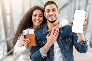 Best Booking App. Happy Arab Couple Holding Passports With Tickets And Blank Smartphone While Posing Together At Airport, Middle Eastern Spouses Recommending New Mobile Application, Mockup photo