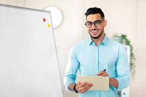 Positive guy English teacher explaining foreign language rules near blackboard, taking notes in clipboard at home office. Young Arab tutor teaching linguistic online course, giving seminar or lecture photo