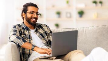 Happy indian guy with glasses working from home, using modern notebook, sitting on couch in cozy living room, copy space. Smiling middle-eastern man freelancer programming or chatting with ladies photo