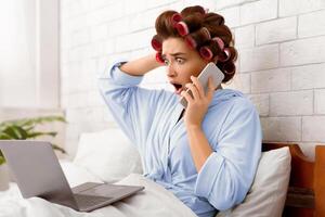 Shocked Housewife Talking On Cellphone Using Laptop Computer Reading Shocking News Online Sitting In Bed Touching Head, Curling Hair With Curlers In Modern Bedroom At Home. Side View Shot photo
