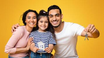 Housing Concept. Happy arab family with little daughter holding paper house and keys, celebrating buying new property, smiling parents and cute female child posing over yellow background, closeup photo
