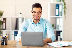 Portrait of confident young Arab guy sitting at his table with laptop computer, typing on keyboard in office. Millennial businessman using portable pc for online job, having virtual meeting photo