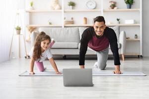 Happy arab father and little daughter having online fitness class, exercising at home together, wearing sportswear, training on yoga mat in front of laptop, enjoying workout, copy space photo