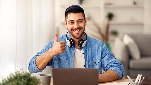 yo como. retrato de contento judío hombre en kippa y auriculares sentado a escritorio utilizando ordenador portátil y demostración pulgares arriba firmar gesto, trabajando o estudiando de forma remota, posando a cámara foto