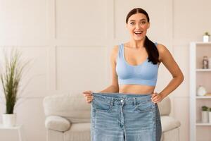Successful Weight Loss. Thin Girl Comparing Old Oversize Jeans After Dieting Standing Indoors. Copy Space photo
