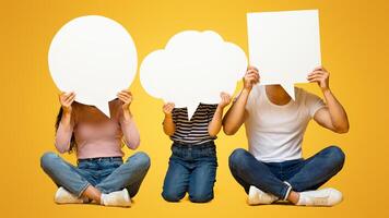 Diverse Thoughts. Father, Mother And Little Daughter Holding Different Empty Speech Bubbles In Front Of Their Faces, Unrecognizable Family Sitting On Floor On Yellow Studio Background, Copy Space photo