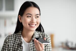 Closeup of smiling asian woman recording voice message on mobile phone while eating at cozy white kitchen, looking at copy space, panorama. Mobile application for communication concept photo