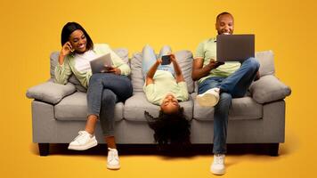 Gadgets addiction. Young black family of three holding and using different electronic devices while sitting on sofa on yellow background. Parents and their daughter with modern gadgets photo