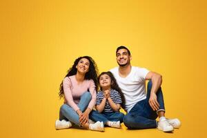 Interesting Offer. Smiling Middle Eastern Family Of Three Looking Up At Copy Space With Excitement, Arab Parents And Daughter Sitting Together Over Yellow Studio Background, Vertical Shot photo