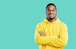 Handsome Black Millennial Man Crossing Hands Posing Smiling To Camera Standing In Studio On Blue Background, Wearing Yellow Hoodie. Shot Of Confident African American Guy photo
