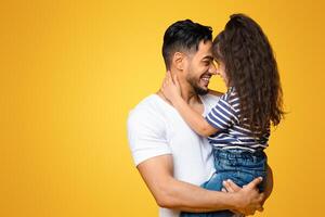 Father Daughter Connection. Portrait Of Happy Middle Eastern Dad Bonding With His Little Daughter, Holding Her On Hands And Touching Noses While Posing Together Over Yellow Background, Copy Space photo