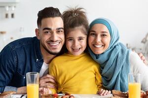 Portrait Of Happy Islamic Parents With Little Daughter Posing In Kitchen While Having Lunch Together, Cheerful Muslim Family Sitting At Table, Eating Tasty Delicious Food And Smiling At Camera photo
