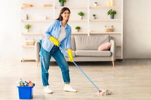 Chores Concept. Portrait of casual woman washing hardwood laminate flooring using string mop pad, standing near bucket with cleaning agents and detergents, posing and looking at camera photo
