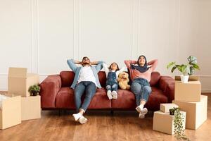 Moving Day. Lovely arab family relaxing on the couch in new home with cardboard boxes around. Happy man, woman in headscarf and little girl lying on sofa and looking up, free copy space photo