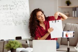 Explaining New Theme. Portrait of young lady tutor having virtual conference, giving online lesson to students using cellphone on tripod. Woman pointing at blank sheet of paper, showing to webcam photo