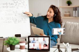Online Lecture Concept. Young lady professor making conference with students, pointing at board, explaining trigonometric functions to students. Woman in headphones filming class on smartphone photo