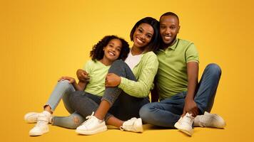 Portrait of loving black parents and their cute daughter smiling together to camera over yellow background. Happy young family of three bonding while sitting on floor in studio photo