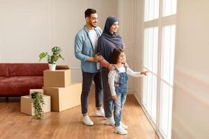 Full Body Length Portrait Of Excited Muslim Family Looking Out And Showing View From Window In Their New Apartment. Renting And House Move Concept. Smiling Little Girl Pointing At Glass photo