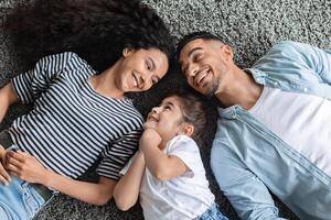 Cheerful arabic family father, mother and preschool daughter laying on floor and laughing, enjoying time together at home. Middle eastern parents spending weekend with their kid, top view photo