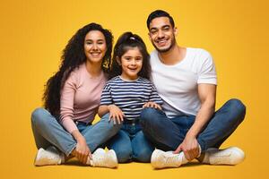 Portrait Of Happy Young Middle Eastern Family Of Three With Little Daughter. Cheerful Arab Parents And Their Cute Female Kid Sitting On Floor Over Yellow Studio Background And Smiling At Camera photo
