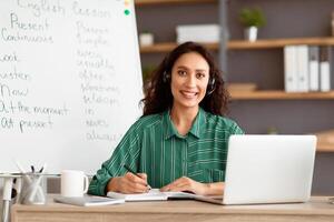Remote Study Concept. Portrait of smiling woman in wireless headset sitting at desk with pc laptop near whiteboard. Happy lady posing and looking at camera, writing, taking notes in paper notebook photo