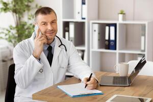 Doctor talking on his smartphone and making notes in a notebook. Medicine professional making phone call with patient and writing in his diary, looking at camera, sitting at desk in his office photo